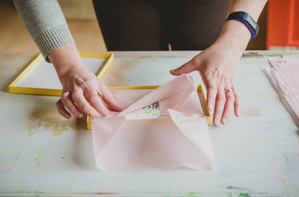 Cards are being folded in pink tissue paper into a yellow box