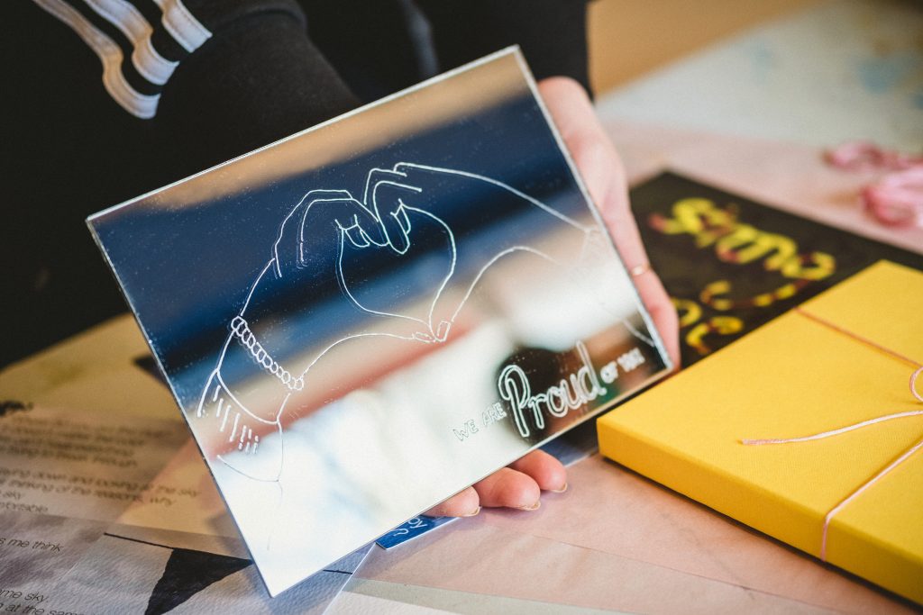 A photograph of someone holding a mirror, onto which is engraved two hands making a loveheart and the phrase 'We are proud of you'