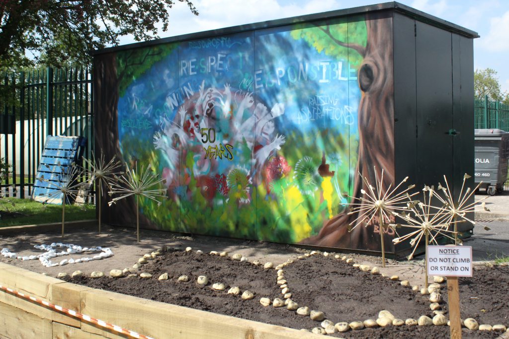 Two trees, hands and leaves are spray painted onto the side of a storage container. Petal shapes are formed on the soil in front, edged by golden stones.