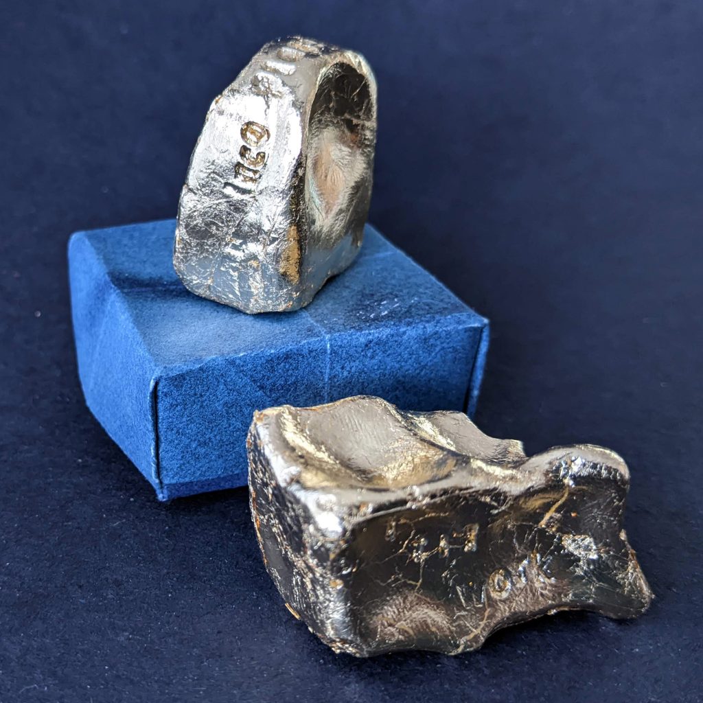 Two gold sculptures, irregularly shaped nuggets. One sits on a blue paper box. 