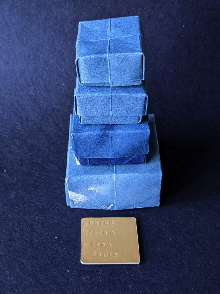 A tower of four, square, blue, folded paper boxes, each one smaller than the one beneath it. A gold card is on the table in front of them imprinted with the words 'Shared Wisdom - Keep Going'