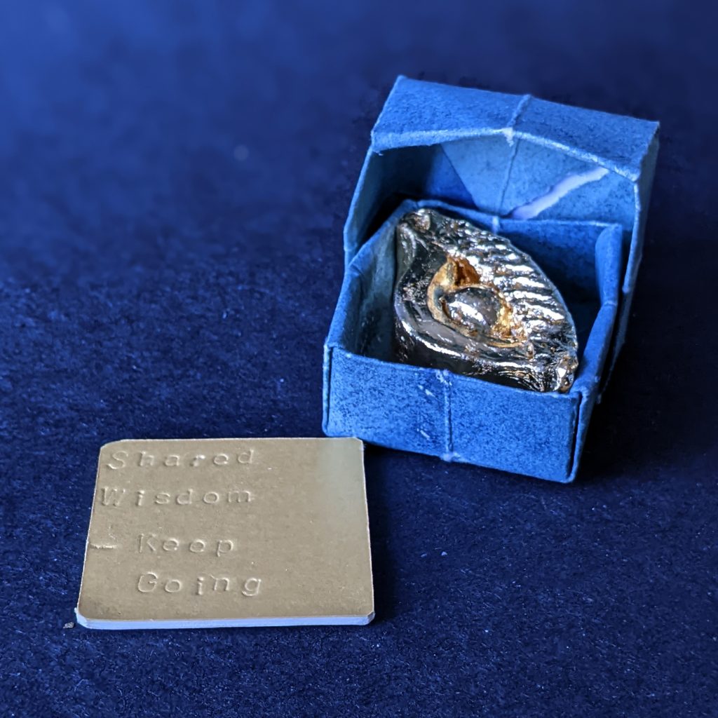 A golden sculpture that looks like a stylised eye sits in a blue paper box. A gold square card is imprinted with the words 'Shared Wisdom - keep going'