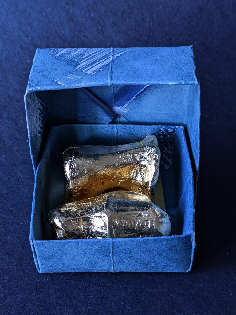 Two irregularly shaped sculptures in gold nestled in a blue paper box whose lid is open.
