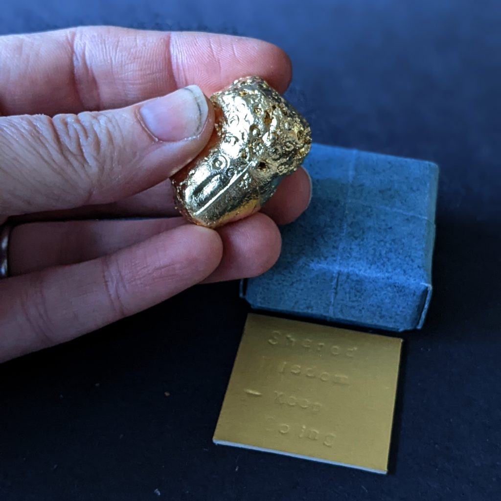 A white-skinned hand holds a golden nugget shaped object that has dimples across its surface. A square, blue paper box sits on the table, with a gold card imprinted with the words 'Shared Wisdom - keep going.'