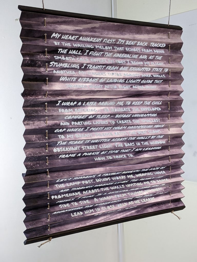 A grey rectangle of concertina-folded paper has the following words cut into it:
My heart awakens first. Its beat back-tracked by the wailing melody that echoes from down the hall. I fight the adrenaline and, as the smooth cold enters my toes I begin my flight. Stumbling, I transit from one exhausted state to another. My fingers tiptoe against dark walls. White ribbons of landing lights guide this journey, night after night after night. 

I wrap a layer around me, to keep the chill from entering - to recreate the enveloped comfort of sleep - before unwrapping and parting layers to create the smallest gap where I press his warm whimpering voice to my breast. Silence rests, the beat slows. The score is written across the 
walls by the observant street light. The bars in the window frame a phrase of time that I am learning how to dance to.

Leafy shadows, a foxtrot beneath the gaze of the lamp post. Winds worry me, weaving under and through wooden limbs. The silhouettes promenade across the walls inviting me to dance freely. I sway. In a repeated two-step, side to side to side.  A warm palm thumps my chest, awakening the knowledge that, for now, I must lead him in close hold as he learns the steps of sleep.