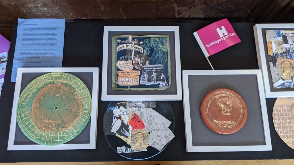 Artwork is laid out on a table, viewed from above. The artworks are circular discs with gold engravings, vinyl records and record sleeves with collaged images over them.