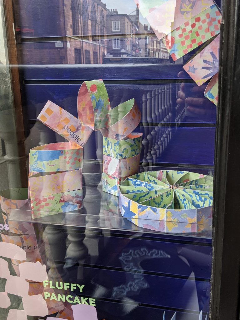 Two sculptures made from coloured paper - one is arch shaped, the other like a wheel laying flat on a shelf. Reflections of the city can be seen in the glass of the cabinet that they are in.