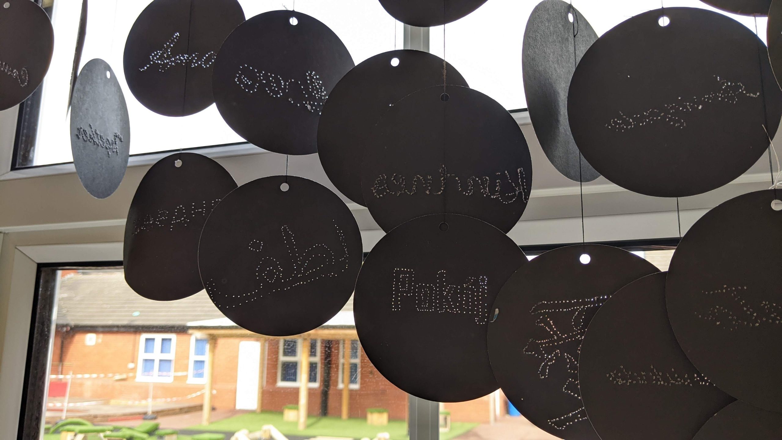 Black circles of card hang in front of a window. Words are written on each card  with tiny pin holes that form the letters. Fragments of words meaning peace, love, and kindness in english, polish, arabic, french and japanese can be seen.