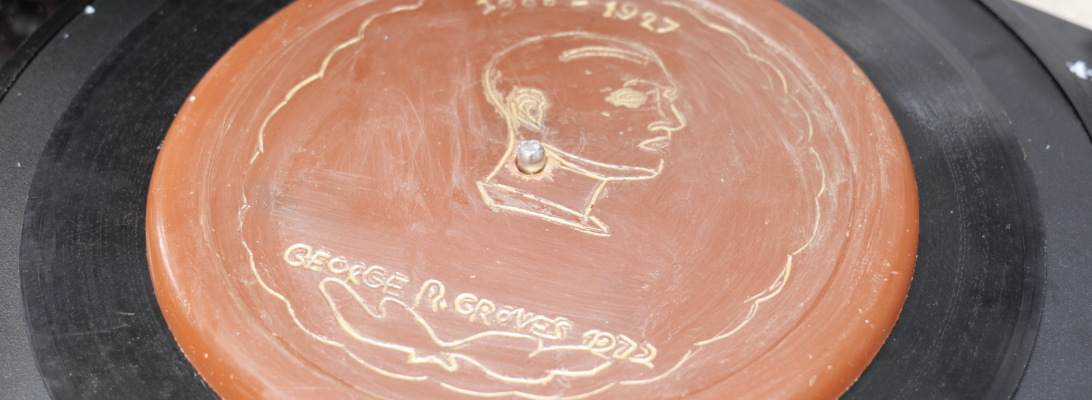 An orange disc made of wax has the profile of a mans face engraved into it. The dates 1888-1927 and the words George R Groves are engraved above and below. The engravings are highlighted in gold.