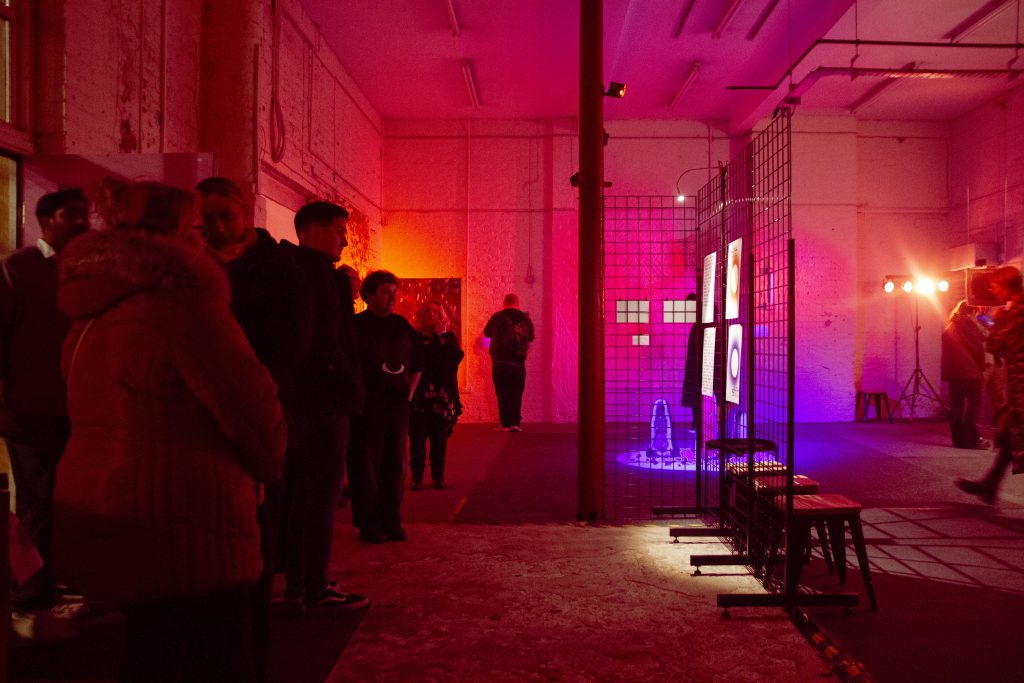 SHIFT Exhibition - an industrial room glows pink and orange. People are looking at artworks at an opening event.