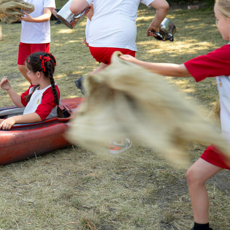 A child is in a canoe on a field. Other children are running past waving sheets of hessian in the air.