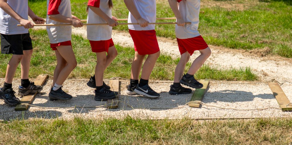 5 children walk across parallel wooden planks whilst carrying rods like they are train pistons
