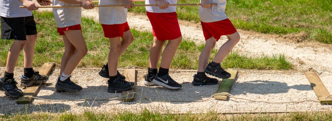 5 children walk across parallel wooden planks whilst carrying rods like they are train pistons